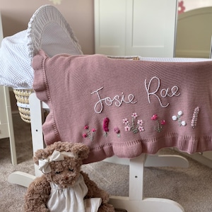 May include: A dusty rose knitted baby blanket with a ruffled edge, featuring the embroidered name Josie Rae in white script and small floral motifs in pink, green, and white. A brown plush teddy bear wearing a white dress sits in front.