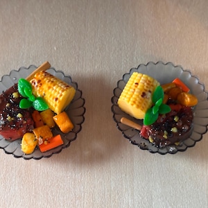 Op de afbeelding: Twee miniatuurborden met eten, elk met gegrilde maïskolf, een stuk biefstuk en diverse groenten. De borden zijn grijs met een geschulpte rand en het eten is zo gerangschikt dat het op een maaltijd lijkt.
