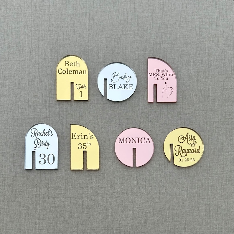 May include: A collection of acrylic place cards in various shapes and colors. The cards feature personalized text, including names, dates, and phrases like "Baby Blake" and "Rachel's Dirty 30." The color palette includes gold, silver, and pink.