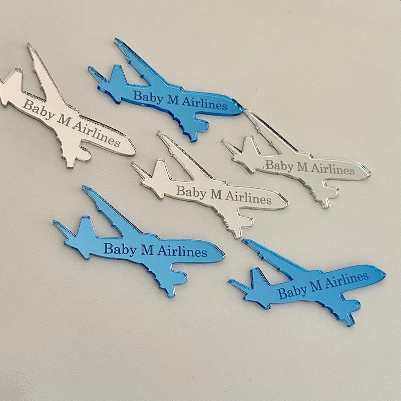 Op de afbeelding: Een verzameling van zes vliegtuigvormige uitsparingen gemaakt van reflecterend materiaal. Drie zijn blauw en drie zijn zilverkleurig. Elk vliegtuigje is voorzien van de tekst Baby M Airlines die in de zijkant is gegraveerd.