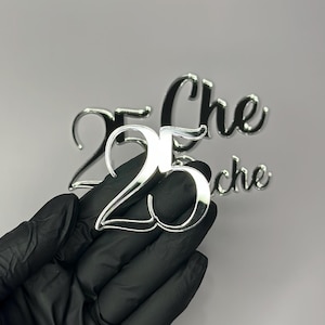 May include: A shiny silver number "25" cake topper held by a gloved hand. The number is in a script font and reflects the light. The word "Che" is also visible in the background, suggesting a celebration or event.