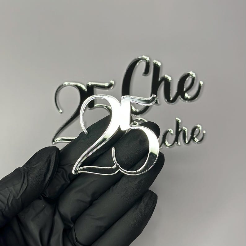 May include: A shiny silver number "25" cake topper held by a gloved hand. The number is in a script font and reflects the light. The word "Che" is also visible in the background, suggesting a celebration or event.