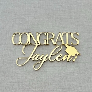 May include: A gold-colored graduation cake topper with the word "CONGRATS" above the name "Jaylen" in cursive script. A small graduation cap is attached to the right side of the name. The topper is made of a shiny material.