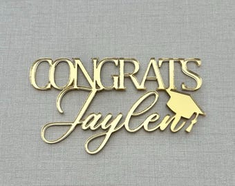 May include: A gold-colored acrylic cake topper featuring the word CONGRATS in a bold, block font above the name Jaylen in a cursive script, with a small graduation cap icon positioned to the right of the name, creating a decorative topper.