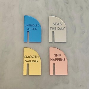 May include: Four colorful, semi-circular nautical-themed decorative pieces. Each piece has a different color: blue, white, gold, and rose gold. Each piece has a different phrase: "UNBRIDLED AT SEA", "SEAS THE DAY", "SMOOTH SAILING", and "SHIP HAPPENS".