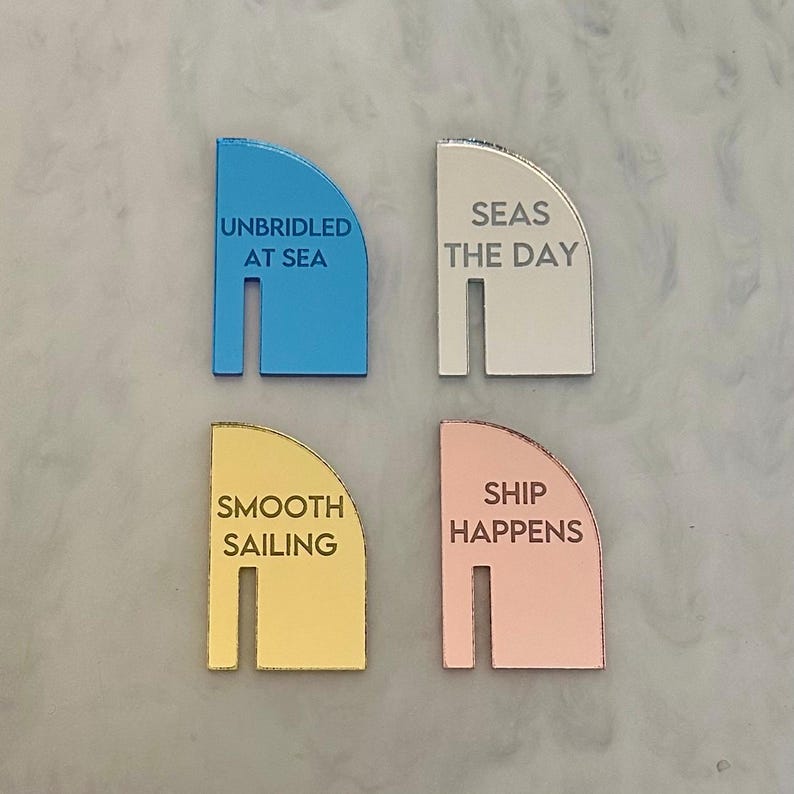 May include: Four colorful, semi-circular nautical-themed decorative pieces. Each piece has a different color: blue, white, gold, and rose gold. Each piece has a different phrase: "UNBRIDLED AT SEA", "SEAS THE DAY", "SMOOTH SAILING", and "SHIP HAPPENS".