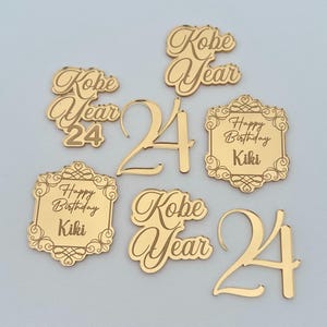 May include: A collection of gold-colored acrylic decorative pieces featuring the text Kobe Year 24, the number 24 in a script font, and rectangular frames with ornate borders containing the text Happy Birthday Kiki in a script typeface.
