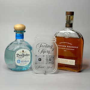 May include: A frosted glass mason jar with the text "Feeling Tipsy?" and two QR codes. The jar is on a white surface with two bottles of alcohol behind it. The bottles are a clear glass bottle with a blue label and a brown glass bottle with a gold label.