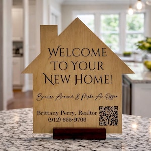 May include: A wooden house-shaped sign with the words "WELCOME TO YOUR NEW HOME!" in elegant script. Below, it reads "Browse Around & Make An Offer." Includes a realtor's name, phone number, and a QR code. The sign is displayed on a countertop.