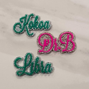 May include: Three glittery name and word charms. The charm "Kokoa" is green with silver glitter. The charm "Dr. B" is pink with silver glitter. The charm "Libra" is green with silver glitter. The charms are on a light gray surface.