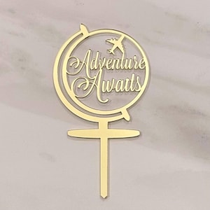 May include: A gold-colored cake topper featuring a globe design with the words "Adventure Awaits" in elegant script. A small airplane is positioned inside the globe. The topper has a long, thin stem for insertion into a cake.