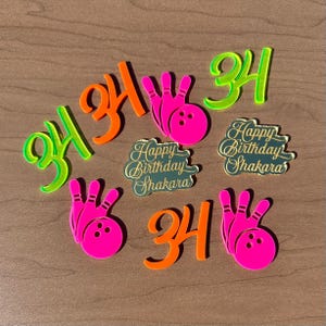May include: A collection of birthday-themed acrylic decorations. The set includes the number "34" in neon orange and green, bowling pin shapes in pink, and two "Happy Birthday Shakara" plaques.