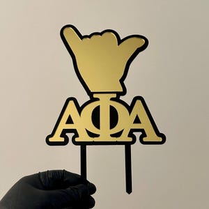 May include: Gold and black cake topper with the letters "AOII" and a gold hand making a shaka sign.