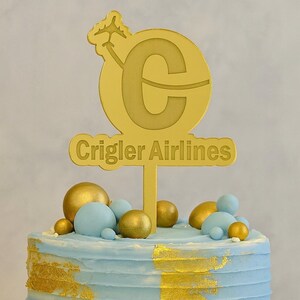 May include: A gold-colored cake topper with the text "Crigler Airlines" and a stylized airplane. The topper is placed on a cake with blue frosting, gold accents, and gold and blue decorative spheres.