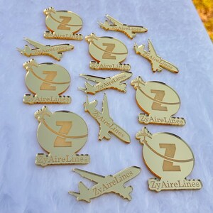 Op de afbeelding: Een verzameling goudkleurige acrylbedels met twee verschillende ontwerpen: een cirkelvormig embleem met een letter Z en een vliegtuigsilhouet, en een aparte vliegtuigvorm. Elk stuk is gegraveerd met de tekst ZyAireLines.