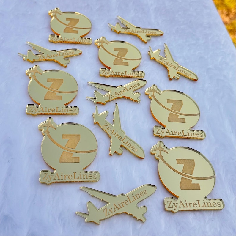 Op de afbeelding: Een verzameling goudkleurige acrylbedels met twee verschillende ontwerpen: een cirkelvormig embleem met een letter Z en een vliegtuigsilhouet, en een aparte vliegtuigvorm. Elk stuk is gegraveerd met de tekst ZyAireLines.