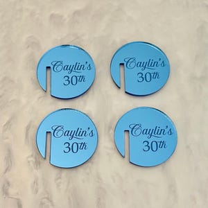 May include: Four light blue, circular coasters with a slot cut out of the side. Each coaster has the text "Caylin's 30th" in a script font. The coasters are arranged on a textured, off-white surface.