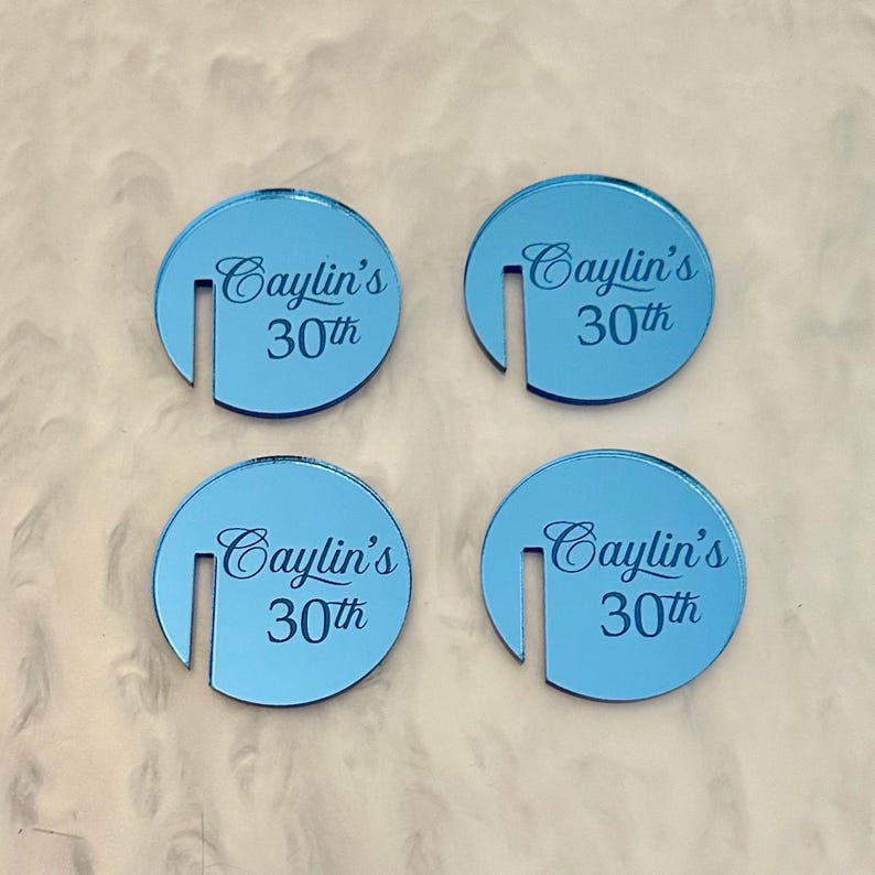 May include: Four light blue, circular coasters with a slot cut out of the side. Each coaster has the text "Caylin's 30th" in a script font. The coasters are arranged on a textured, off-white surface.