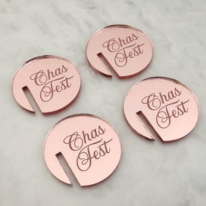 May include: Four rose gold drink charms with a slot for a stem. Each charm has the text "Chas Fest" in a cursive font. The charms are circular and have a reflective surface.