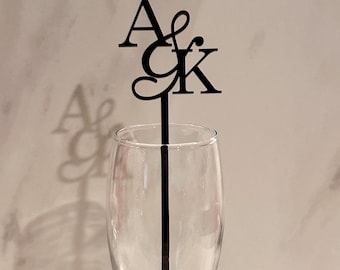 May include: Black acrylic cake topper with the letters "A & K" in a decorative font. The topper is inserted into a clear champagne flute, with the shadow of the letters visible on the background.