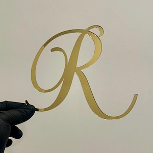 May include: A gold mirror acrylic letter R in a cursive font. The letter is cut out and has a shiny, reflective surface.