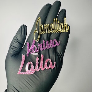 May include: Three custom name cutouts in gold, purple glitter, and pink glitter. The names are "Jamelliah", "Karissa", and "Laila". The cutouts are held in a black gloved hand against a white background.