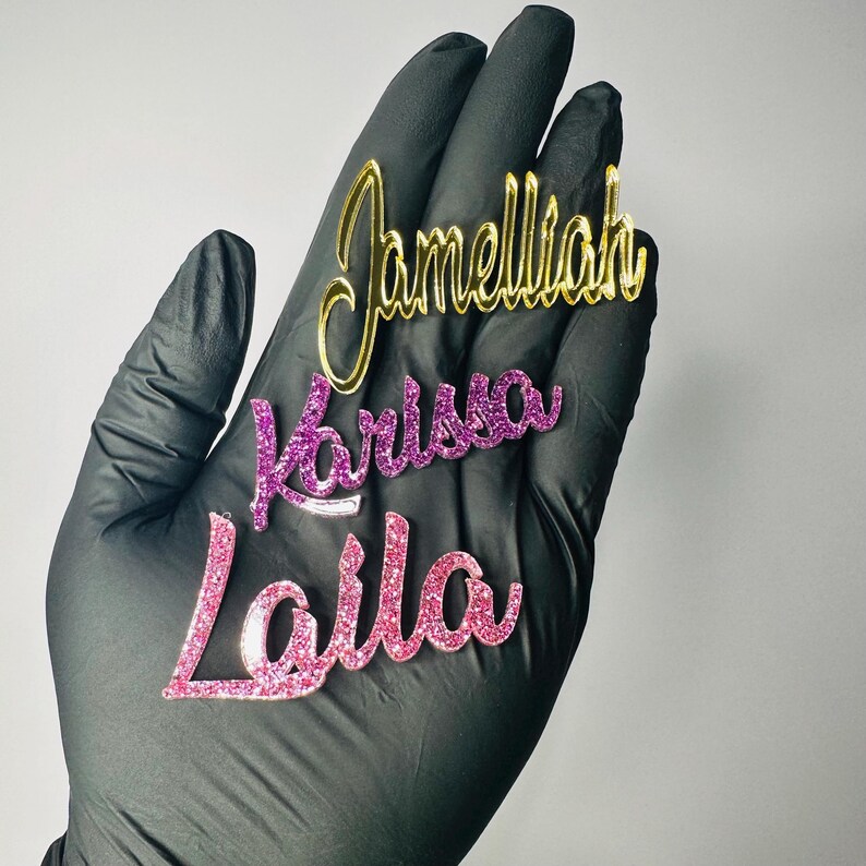 May include: Three custom name cutouts in gold, purple glitter, and pink glitter. The names are "Jamelliah", "Karissa", and "Laila". The cutouts are held in a black gloved hand against a white background.