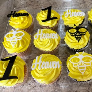 May include: A dozen cupcakes with yellow frosting, decorated with black number one toppers, white bee-shaped toppers, and white toppers that spell "Heaven". The cupcakes are in a clear plastic container.