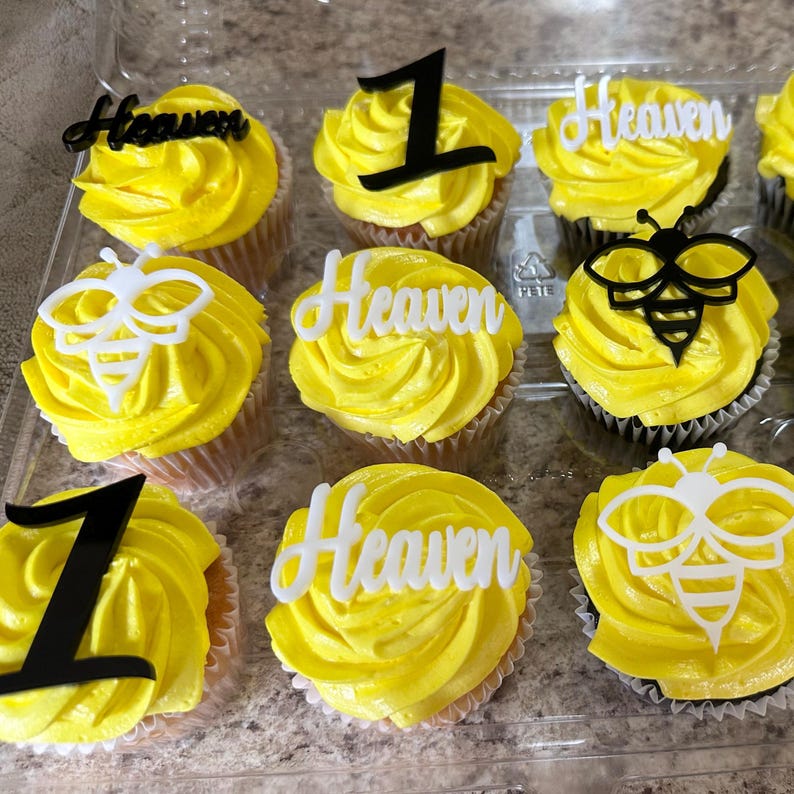 May include: A dozen cupcakes with yellow frosting, decorated with black number one toppers, white bee-shaped toppers, and white toppers that spell "Heaven". The cupcakes are in a clear plastic container.