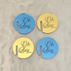 May include: Four circular, decorative coasters. Two are blue, two are gold. Each coaster has a slot cut out of the side and the words "Oh Boy" in a script font. The coasters are likely for a baby shower or gender reveal.