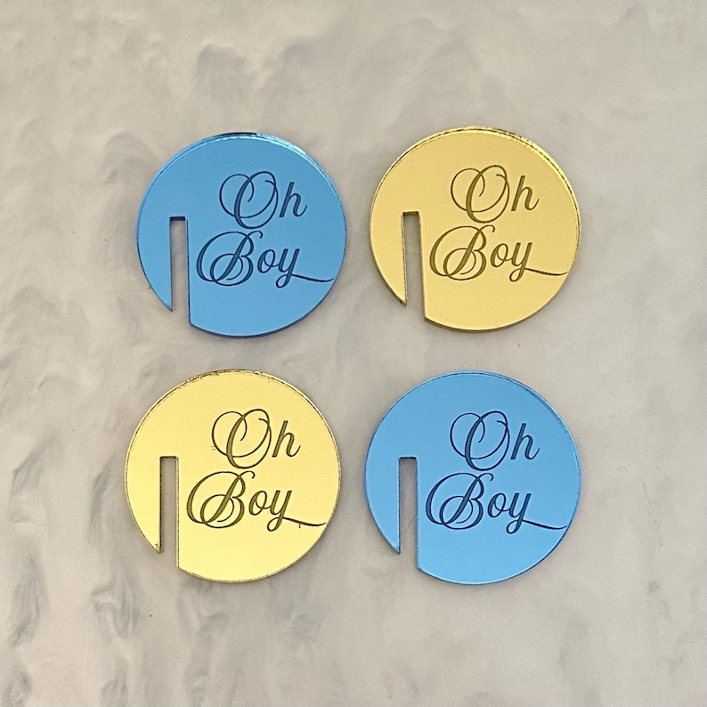 May include: Four circular, decorative coasters. Two are blue, two are gold. Each coaster has a slot cut out of the side and the words "Oh Boy" in a script font. The coasters are likely for a baby shower or gender reveal.