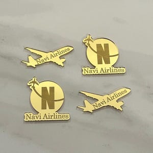 Op de afbeelding: Vier goudkleurige reflecterende acrylstukken met luchtvaartontwerpen. Twee stukken zijn vliegtuigsilhouetten met de tekst Navi Airlines, en twee zijn cirkelvormige emblemen met een grote letter N, een klein vliegtuig en de tekst Navi Airlines.
