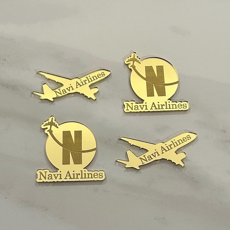 Op de afbeelding: Vier goudkleurige reflecterende acrylstukken met luchtvaartontwerpen. Twee stukken zijn vliegtuigsilhouetten met de tekst Navi Airlines, en twee zijn cirkelvormige emblemen met een grote letter N, een klein vliegtuig en de tekst Navi Airlines.
