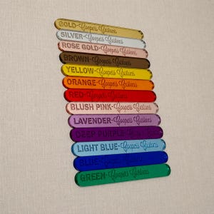 Op de afbeelding: Een stapel van twaalf rechthoekige acryl kleurstalen, elk gegraveerd met de kleurnaam en de tekst Coopers Customs. De kleuren zijn goud, zilver, ros&eacute;goud, bruin, geel, oranje, rood, oudroze, lavendel, donkerpaars, lichtblauw, blauw en groen.