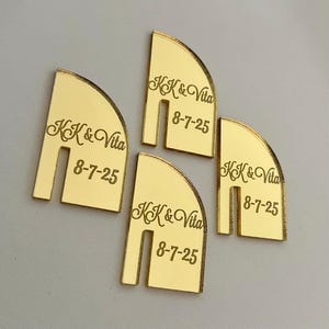 May include: Four gold-colored, semi-circular wedding place cards. Each card has a slot cut out at the bottom and features the names "KK & Vita" and the date "8-7-25" engraved in a script font.