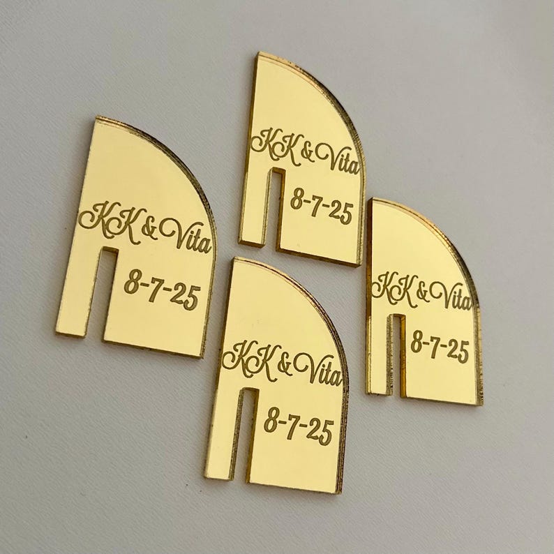 May include: Four gold-colored, semi-circular wedding place cards. Each card has a slot cut out at the bottom and features the names "KK & Vita" and the date "8-7-25" engraved in a script font.