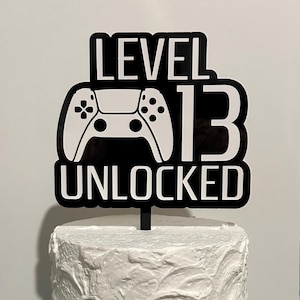 May include: Black and white cake topper with a video game controller graphic and the text "Level 13 Unlocked".