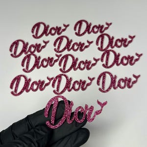 May include: Pink glitter "Dior" cutouts in a cursive font. The word is repeated multiple times. One cutout is held by a gloved hand. The cutouts are likely for crafting or decoration.
