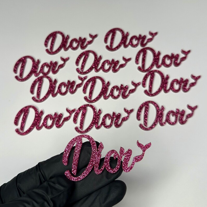 May include: Pink glitter "Dior" cutouts in a cursive font. The word is repeated multiple times. One cutout is held by a gloved hand. The cutouts are likely for crafting or decoration.