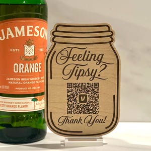May include: A light-colored wooden sign shaped like a mason jar featuring the engraved text Feeling Tipsy? and Thank You! with a central QR code. Beside it stands a green glass bottle of Jameson Orange Irish whiskey with an orange label.