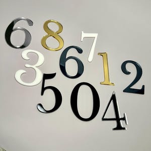 May include: Assortment of acrylic numbers in various colors, including black, white, gold, and silver. The numbers include 0, 1, 2, 3, 4, 5, 6, 7, and 8.