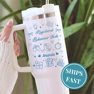 May include: White tumbler with a handle, featuring the text "Registered Behavior Tech" and the name "Amanda" in blue script. The design includes various blue icons. A teal circle with the words "SHIPS FAST" is on the bottom.