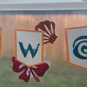 May include: A decorative banner spelling "TWO" with glittery blue letters on white pennants with orange trim. The banner includes tropical-themed cutouts of a leaf, seashell, flower, and a sailboat, strung together with twine.
