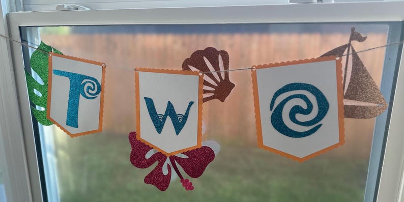 May include: A decorative banner spelling "TWO" with glittery blue letters on white pennants with orange trim. The banner includes tropical-themed cutouts of a leaf, seashell, flower, and a sailboat, strung together with twine.