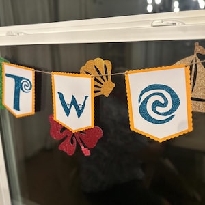 May include: A banner spelling "TWO" on white pennants with orange scalloped edges. The letters and symbols are in blue glitter. Additional cutouts include a gold seashell, a red hibiscus flower, and a gold sailboat.