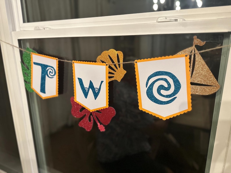 May include: A banner spelling "TWO" on white pennants with orange scalloped edges. The letters and symbols are in blue glitter. Additional cutouts include a gold seashell, a red hibiscus flower, and a gold sailboat.