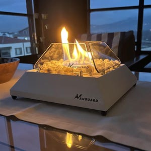 May include: A white tabletop fireplace with a glass firebox and a flame burning inside. The fireplace is labelled "Vanguard" and is sitting on a glass table.