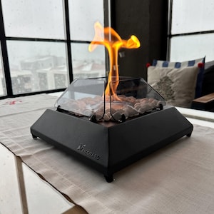 May include: A black tabletop fireplace with a glass firebox and a flame burning inside. The fireplace is on a white table runner and has the word "Vanguard" printed on the front.