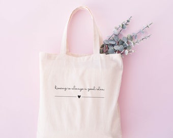 May include: A light beige canvas tote bag with two handles, featuring the black printed text "Kissing is always a good idea" above a horizontal line with a small heart in the center. A sprig of green eucalyptus leaves is tucked inside the bag.