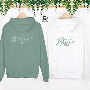 May include: Two hooded sweatshirts, one green and one white, with the words "Bride" and "Bridesmaid" on the back. The date "28.6.24" is printed below each word.